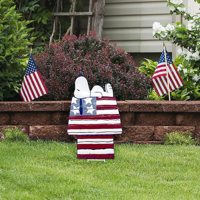 This is a lifestyle image showing this decorative garden stake in a garden. This decorative garden stake is designed to look like the famous Peanuts character, Snoopy. Snoopy is sleeping on top of his doghouse and the house is designed to look like the American flag: red, white and blue stars and stripes.