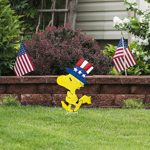 This is a lifestyle image showing a garden stake shaped like the Peanuts character Woodstock. He's wearing a red, white and blue top hat.