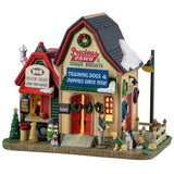 This is a Lemax lighted building designed to look like a a barn that's been turned into a pet store. There's snow on the roof and decorated Christmas trees and wreaths. There's a booth to the left with signs that read "DOGGIE TREATS FOR TOP DOGS!" and "FLAVORS: BEEF, PORK, CHICKEN." There are dogs waiting for treats and dogs being trained off to the right. There are signs above the front door that read "PRECIOUS PAWS & DOGGIE BISCUITS" and "TRAINING DOGS & PUPPIES SINCE 1958."