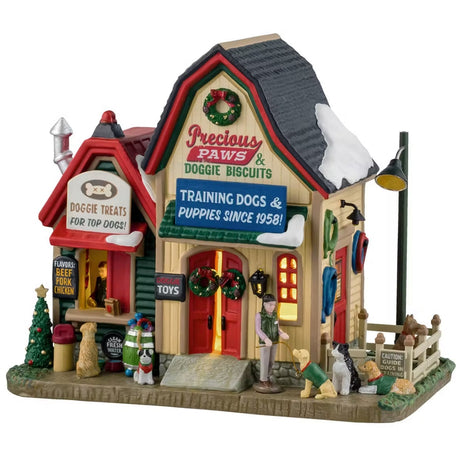 This is a Lemax lighted building designed to look like a a barn that's been turned into a pet store. There's snow on the roof and decorated Christmas trees and wreaths. There's a booth to the left with signs that read "DOGGIE TREATS FOR TOP DOGS!" and "FLAVORS: BEEF, PORK, CHICKEN." There are dogs waiting for treats and dogs being trained off to the right. There are signs above the front door that read "PRECIOUS PAWS & DOGGIE BISCUITS" and "TRAINING DOGS & PUPPIES SINCE 1958."