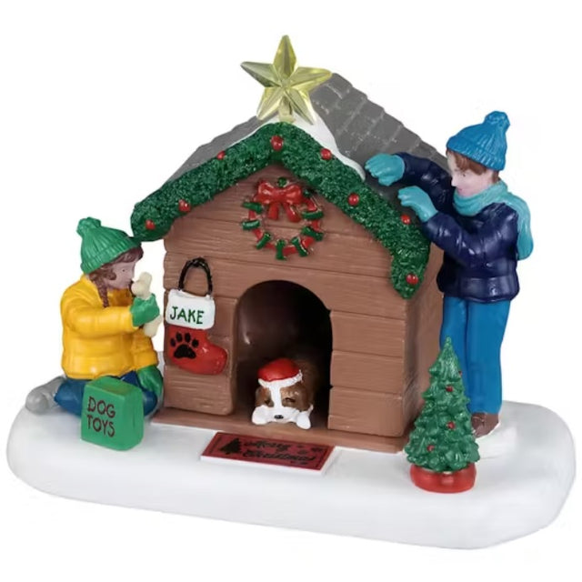 This is a Lemax table accent designed to look like a wooden dog house with a dog laying down inside. He's wearing a Santa hat. There's a young boy and young girl decorating the doghouse with garland, a wreath, a star, a stocking and a small decorated Christmas tree. The girl has a bag full of dog toys.
