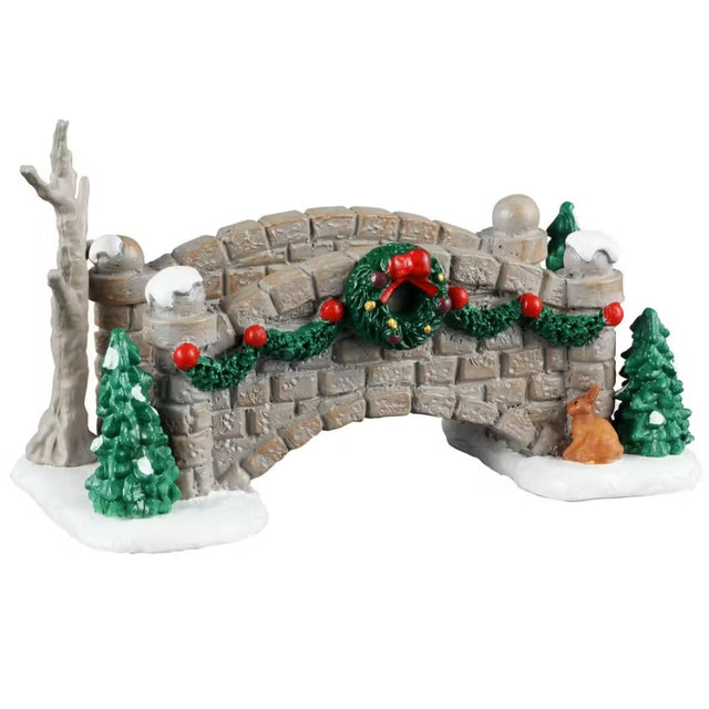 This is a Lemax village accessory designed to look like a gray brick bridge covered in snow. On one side, there's a bare tree and a short pine tree. On the other side, there are two short pine trees. The front of the bridge is covered in green garland and a decorated wreath. At the base of the bridge, there's a little orange bunny.
