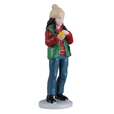 This person is wearing a tan colored winter hat, a red long sleeve shirt, a green vest, blue jeans and black shoes. She's holding food in her hand.
