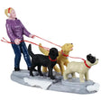 This image shows a Lemax figurine designed to look like a girl wearing a purple sweater and red earmuffs walking three dogs on red leashes. One dog is black, one dog is yellow and one dog is white with black ears. There's snow on the ground.