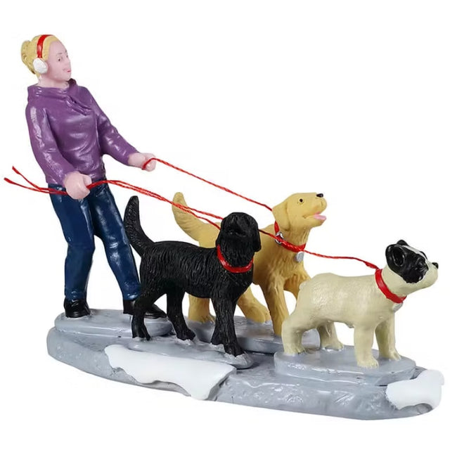 This image shows a Lemax figurine designed to look like a girl wearing a purple sweater and red earmuffs walking three dogs on red leashes. One dog is black, one dog is yellow and one dog is white with black ears. There's snow on the ground.