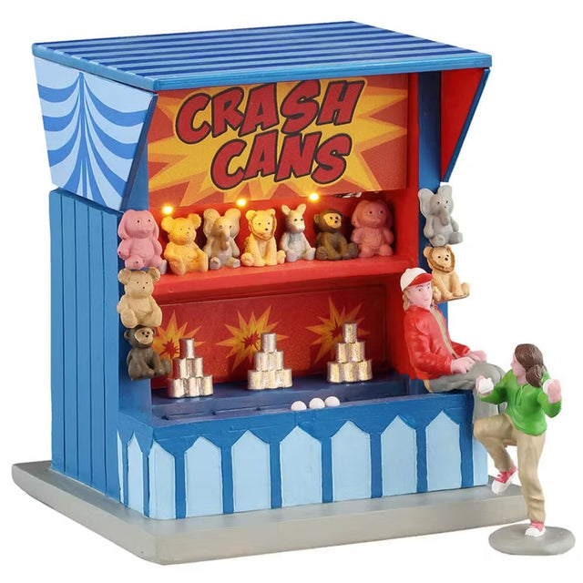 This is a Lemax table accent that is designed to look like a blue carnival booth. It reads "CRASH CANS" at the top of the booth. Underneath the sign, there's a shelf full of stuffed animal prizes. There are silver cans stacked up on another shelf. A girl wearing a green sweatshirt is gearing up to throw a ball at the cans as the booth worker watches.