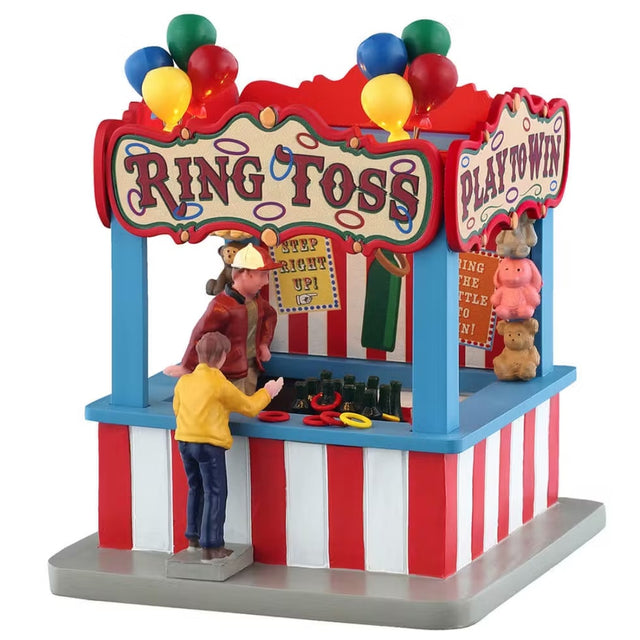 This is a Lemax table accent designed to look like a Ring Toss booth at a carnival. There are red, green, blue and yellow balloons on the roof. There are teddy bear prizes on the back wall of the booth. There's a man inside watching a young boy throw rings on the bottles. One of the rings has landed on the bottle.