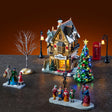 This is a lifestyle image showing the set on display. There's the building with a lamp post on either side. There's also the telephone booth on the right side and a tree on either side. In front, there's a table piece showing three people in Victorian clothes shopping and four Victorian carolers singing for another couple.