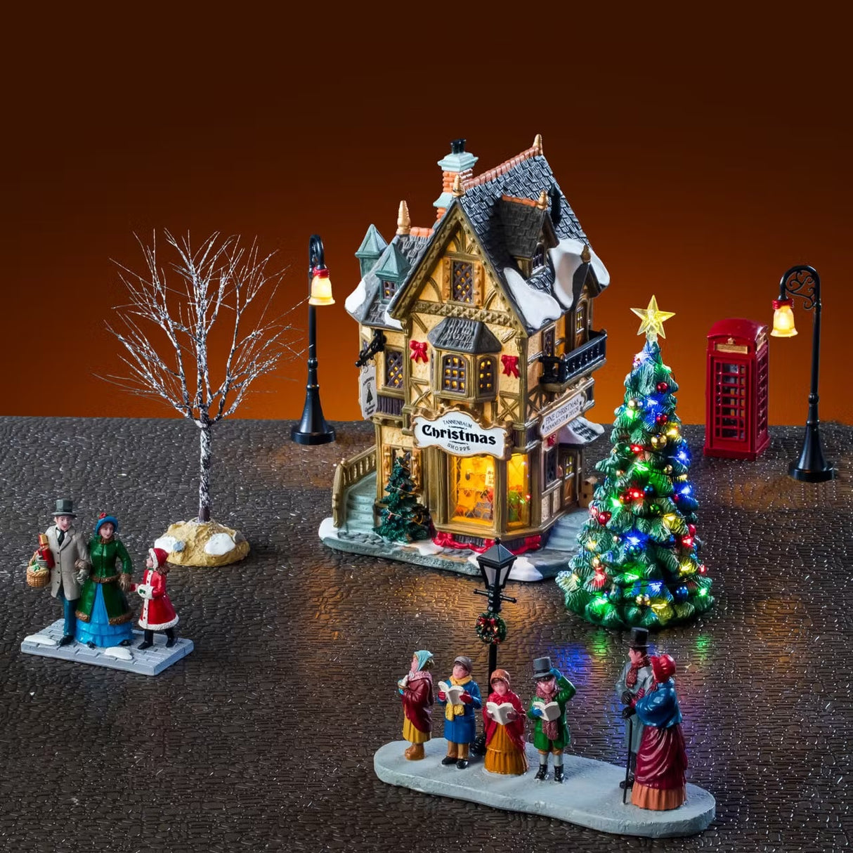 This is a lifestyle image showing the set on display. There's the building with a lamp post on either side. There's also the telephone booth on the right side and a tree on either side. In front, there's a table piece showing three people in Victorian clothes shopping and four Victorian carolers singing for another couple.