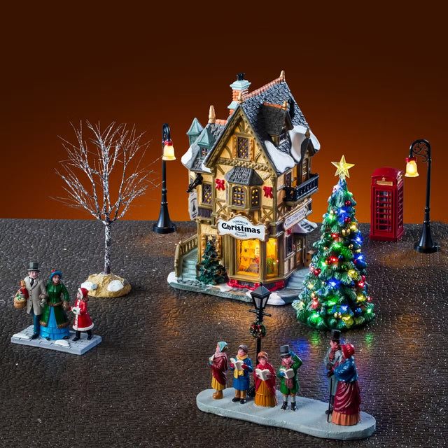 This is a lifestyle image showing the set on display. There's the building with a lamp post on either side. There's also the telephone booth on the right side and a tree on either side. In front, there's a table piece showing three people in Victorian clothes shopping and four Victorian carolers singing for another couple.