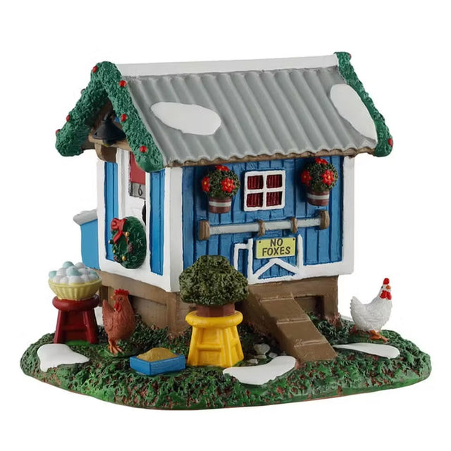 This is a Lemax table accent designed to look like a blue chicken coop. The roof is decorated with garland and the sides are decorated with wreaths and other flowers. There's a sign that reads "NO FOXES." There are two chickens walking around in the grass and snow. There's a bowl of eggs.