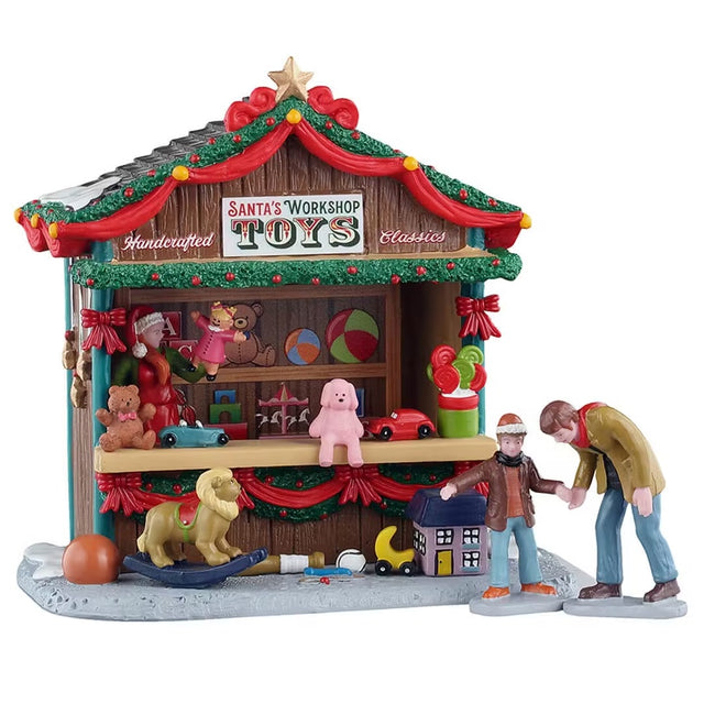 This is a Lemax table piece designed to look like a wooden booth full of toys. It's decorated with red ribbon and garland. There's a gold star on the roof. Some of the toys include stuffed animals, a rocking horse, tiny cars, beach balls, blocks and more. A man and his son are deciding which toys they want.