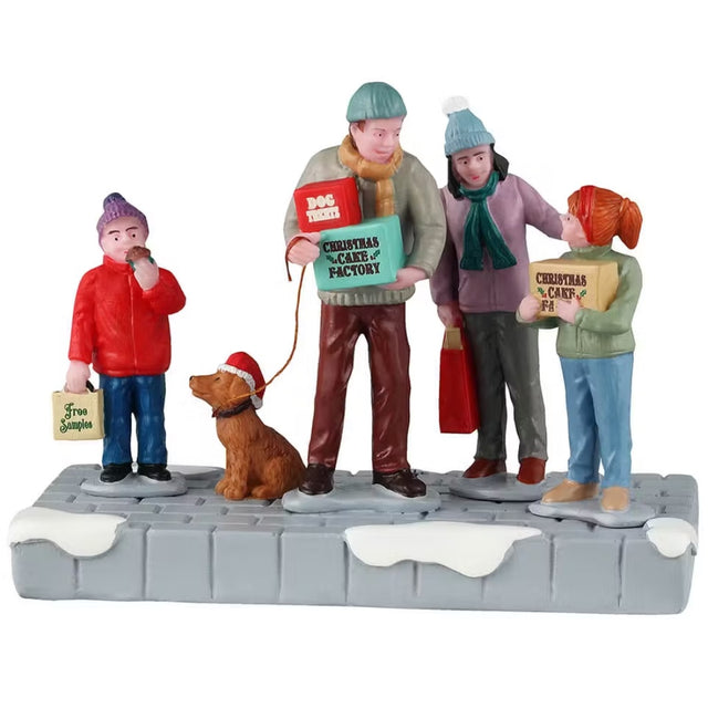 This Lemax table accent is designed to look like a family of four wearing coats, winter hats and scarves standing on a snowy sidewalk. They're all holding boxes. The young son is holding a box that reads "FREE SAMPLES." The dad is holding two boxes: "Dog Treats" and "Christmas Cake Factory." The mom is holding a red bag. The young daughter is holding a box that reads "Christmas Cake Factory." The dad is also holding the leash of their dog who is wearing a Santa hat.