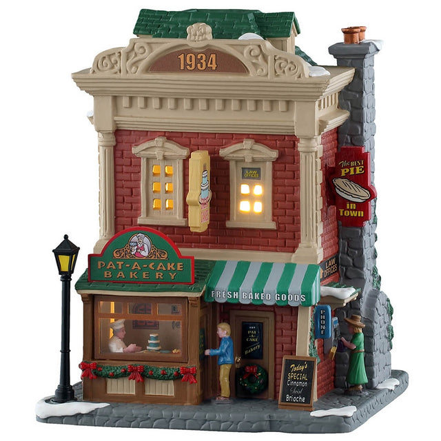 This is a Lemax lighted building designed to look like a tall, red brick building turned into a bakery. There's a lamp post on the sidewalk, along with snow. There's a man about to walk into the building and a woman reading the menu on the side of the building. Inside the window, you can see the chef putting icing on a cake.
