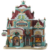This is a Lemax lighted building designed to look like a large mansion turned into a pastry and coffee shop. There's snow on the roof and on the sidewalk. There are lots of teal colored accents. There's a wreath above the window and garland above the door. There are two windows you can see into: one of them shows two people at a table looking at a menu and the other you can see a woman behind the counter.