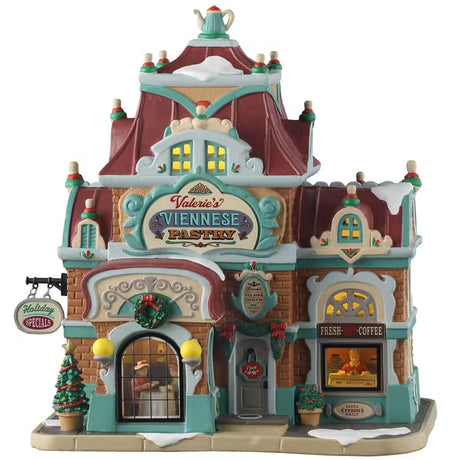 This is a Lemax lighted building designed to look like a large mansion turned into a pastry and coffee shop. There's snow on the roof and on the sidewalk. There are lots of teal colored accents. There's a wreath above the window and garland above the door. There are two windows you can see into: one of them shows two people at a table looking at a menu and the other you can see a woman behind the counter.