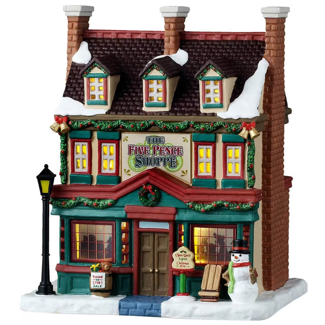 This is a Lemax lighted building designed to look like a tall old timey cottage turned into a shop. There's snow on the roof and on the sidewalk. It's decorated with garland, wreaths and red ribbons. There's a lamp post on the left and a cute snowman on the right.
