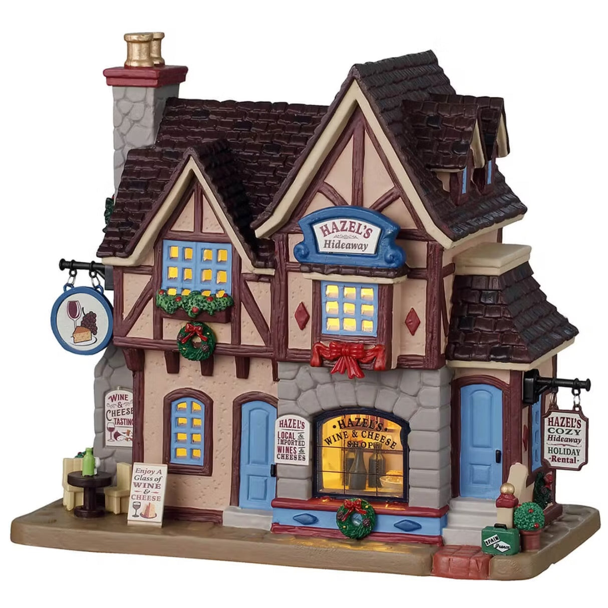 This is a Lemax lighted building designed to look like an old timey cottage turned into a holiday rental / wine and cheese shop. It's decorated with wreaths and red ribbon. There are signs that read "WINE & CHEESE TASTING," "ENJOY A GLASS OF WINE & CHEESE," "HAZEL'S LOCAL IMPORTED WINES & CHEESES" and "HAZEL'S COZY HIDEAWAY HOLIDAY RENTAL."
