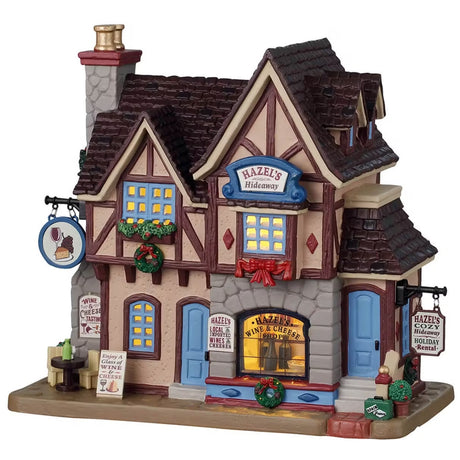 This is a Lemax lighted building designed to look like an old timey cottage turned into a holiday rental / wine and cheese shop. It's decorated with wreaths and red ribbon. There are signs that read "WINE & CHEESE TASTING," "ENJOY A GLASS OF WINE & CHEESE," "HAZEL'S LOCAL IMPORTED WINES & CHEESES" and "HAZEL'S COZY HIDEAWAY HOLIDAY RENTAL."