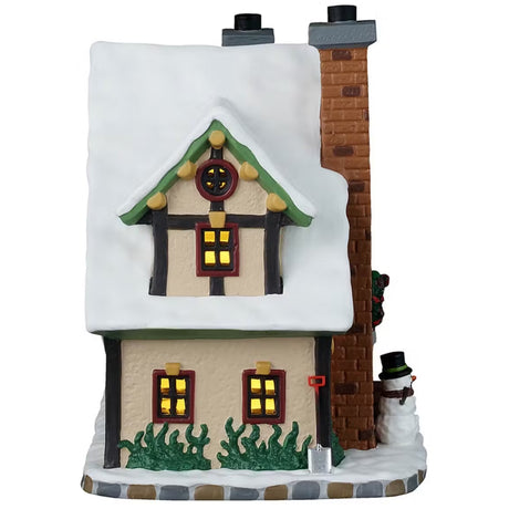 This is the side of the building. There are four windows with light shining through. There's grass peeking out from the snow and a silver shovel leaning up against the building.