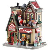 This is a Lemax lighted building designed to look like a tall, brick shop. On top near the roof, there are four statues: one is a dog dressed as Santa, one is a cat dressed as a reindeer, one is a dog wearing a tuxedo and top hat and one is a cat wearing a pink sweater. There are pets dressed up on the sidewalk: one is a cat wearing a hat and a red bandana, one is a dog wearing a polka dot sweater, one is a dog dressed as a ballerina and one is a dog wearing reindeer ears.