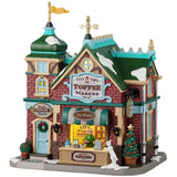 This is a Lemax lighted building designed to look like a red brick store. There's a sign over the front door that reads "MADE FRESH DAILY." There's a wreath on the front door and two potted trees. There's a booth in the front you can walk up to and order candy from the person at the register. There's a white dog peeking over.