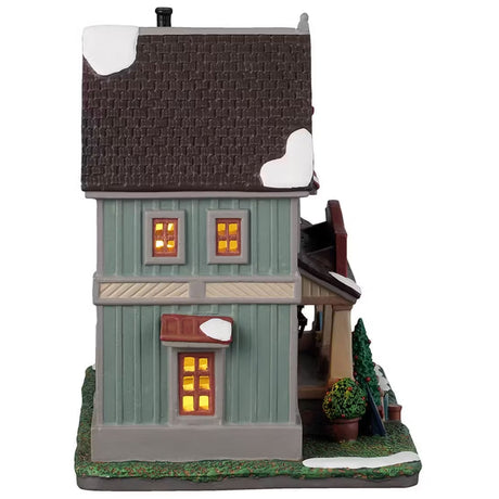 This is the side of the building. There's snow on the roof and on the front lawn. There are three windows on the side with lights shining through.