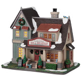 This is a Lemax lighted building. There's a sign above the front door that reads "WADDLETON'S BED & BREAKFAST." There's a wreath near the roof and a wreath on the front door. There are decorated potted trees on either side of the front stairs. There's a sign on the front lawn that reads "WELCOME TO OUR ANNUAL HOLIDAY WRITER'S RETREAT. FRIDAY THROUGH SUNDAY."