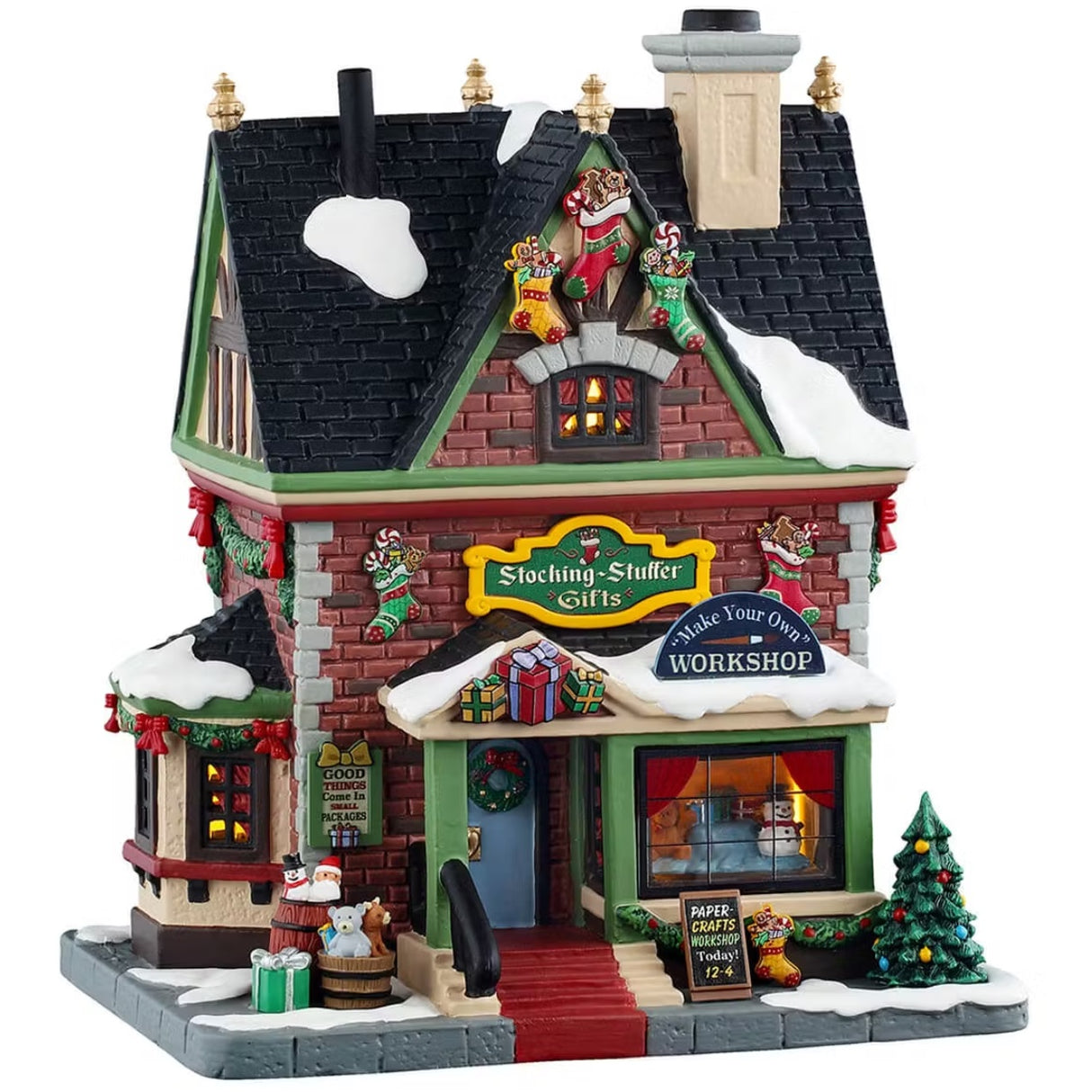 This is a Lemax lighted building designed to look like a tall, red, brick building. All over, there are stocking decorations with gingerbread cookies and peppermint candies sticking out. There's a window display of a snowman inside. There are wreaths and garlands and decorated trees. There are barrels with stuffed animals inside and a present on the sidewalk.