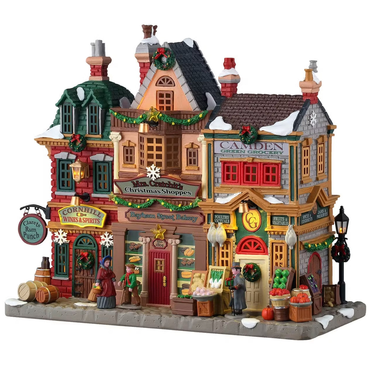 This is a Lemax lighted building designed to look like a strip of shops. From left to right the shops are: Cornhill Wines & Spirits, Bayham Street Bakery and Camden Green Grocery. There's snow on the roof and on the sidewalk. There are barrels and crates of fruits and vegetables along the sidewalk.