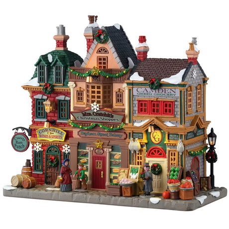 This is a Lemax lighted building designed to look like a strip of shops. From left to right the shops are: Cornhill Wines & Spirits, Bayham Street Bakery and Camden Green Grocery. There's snow on the roof and on the sidewalk. There are barrels and crates of fruits and vegetables along the sidewalk.