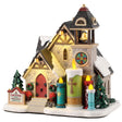 This is a Lemax lighted building designed to look like a stone church. There's a bell tower and a circular glass-stained window. In front of the church, there are four large candle statues. There's snow on the roof and on the ground.