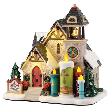 This is a Lemax lighted building designed to look like a stone church. There's a bell tower and a circular glass-stained window. In front of the church, there are four large candle statues. There's snow on the roof and on the ground.