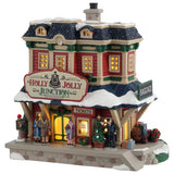 This is a Lemax lighted building designed to look like an old Victorian-era style building turned into a train station. There are people standing outside waiting for their trains. They're wearing winter clothes. There's a bellhop waving beside a few pieces of luggage. There are decorated Christmas trees and wreaths. There's snow on the roof and on the sidewalk.