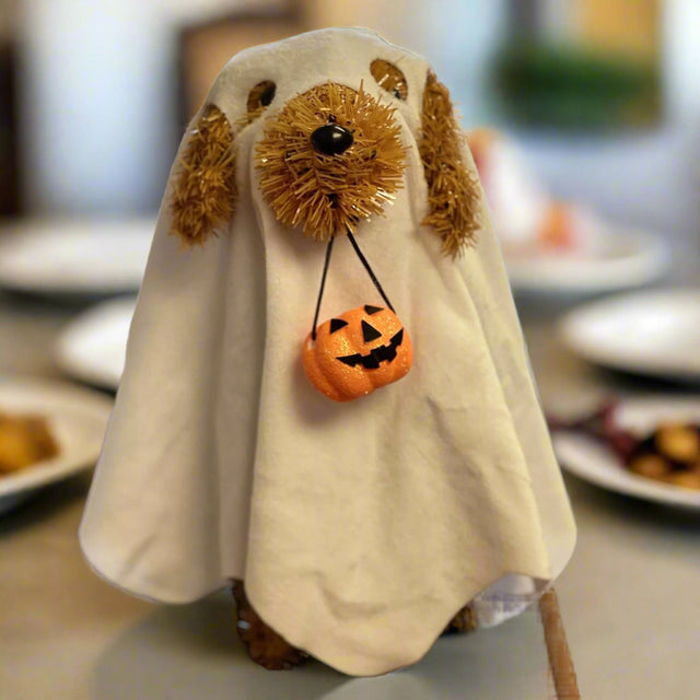 This tabletop decoration is a brown tinsel dog with black eyes and a black nose. He's wearing a soft, white sheet with eyeholes and a hole cut for his snout - so he looks like a ghost! He's holding a pail in his mouth that's shaped like a jack-o-lantern.