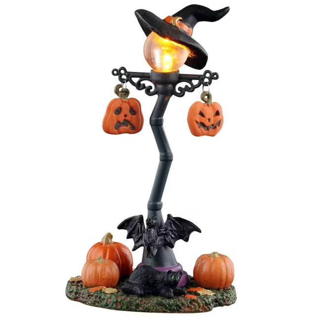 This is a Lemax Spooky Town lamp post decorated with a witch hat, two hanging jack-o-lanterns and a bat at the base. Underneath the lamp there are three pumpkins and one black cat with green eyes.