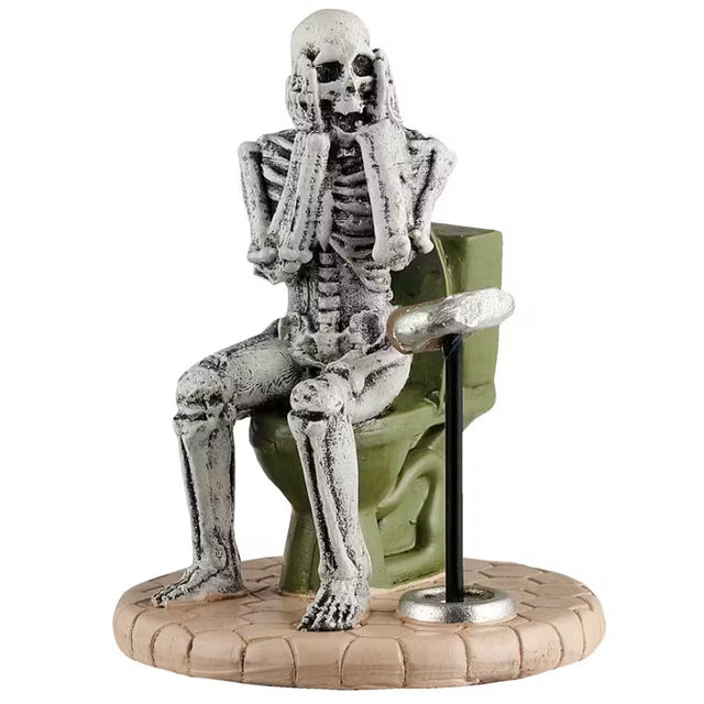 This is a Lemax Spooky Town figurine that's designed to look like a skeleton sitting on a green toilet. With his hands on his face, shocked, he's looking at the toilet paper holder and realizing there's no toilet paper left.