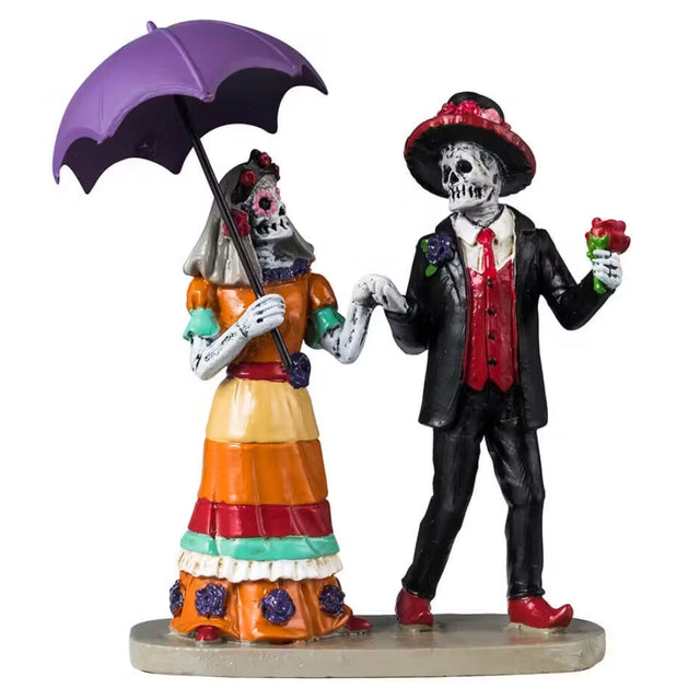 This image shows a Lemax Spooky Town figurine designed to look like two skeletons dressed up for Dia de los Muertos. The skeleton on the left is a woman wearing an orange, teal, red, yellow and purple striped dress and is holding a purple umbrella. The skeleton of the right is a man wearing a black suit with a red vest and red shoes. He's holding a red rose in his hand.