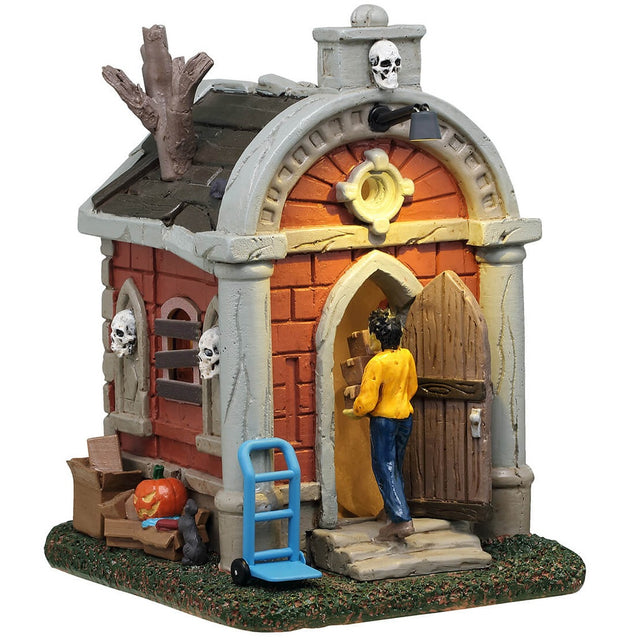 This is a Lemax Spooky Town building designed to look like a mausoleum. The windows are boarded up, there's a skull above the front door and along the sides of the building. A man is carrying boxes into the building and there are boxes along the side. There's a blue furniture mover with wheels near the front steps.