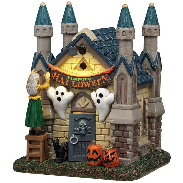 This is a Lemax Spooky Town building designed to look like a small house that's being decorated for Halloween. There's a woman standing on a ladder hanging up a banner that reads "HAPPY HALLOWEEN." There are two ghosts put up on either side of the front door and two jack-o-lanterns on the ground. The door has a skull on it and the door is open with a black cat peeking out.
