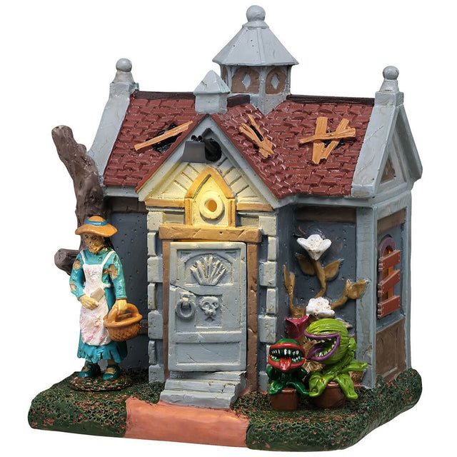 This is a Lemax Spooky Town building designed to look like a rundown garden shed. There are holes patched up with slats of wood, windows boarded up and plants with teeth growing near the front door. A woman wearing a blue dress and a white apron is standing near the front door holding a basket.