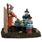 This is a different angle of the Lemax Spooky Town table accent designed to look like three witches sitting around stirring their individual cauldrons. There's a white board that reads "TOAD SOUP. Ingredients: 3 toads, 1 cup of swamp water, 1 cup of flies and 1 cup of worms." One witch is sitting on a stool and is wearing a blue cloak with a black hat. One witch is sitting on a tree stump and is wearing a dark blue cloak with a black hat. One witch is standing and is wearing an orange cloak with a pink hat.