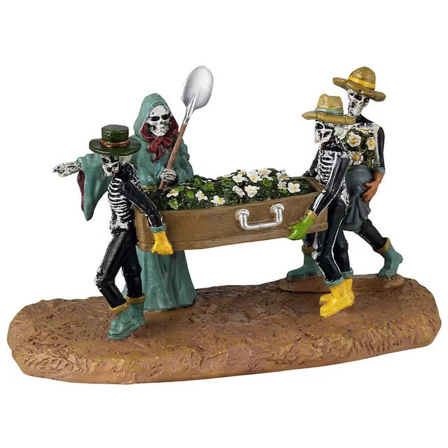 This is a Lemax Spooky Town table accent designed to look like three skeletons carrying a coffin full of daisies. They're all wearing garden hats and rubber boots. There's a Grim Reaper wearing a teal cloak pointing the way.
