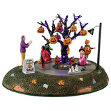 This Lemax Spooky Town table accent is designed to look like a bunch of kids wearing Halloween costumes in a parade around a spooky tree decorated with jack-o-lanterns. There's a parent off to the side taking pictures. One kid is dressed like Superman, one is dressed like a witch and she's walking a dog, one kid is dressed like a pumpkin, one kid is dressed like a piece of candy corn and the other kid is dressed like a cat.