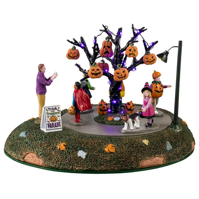 This Lemax Spooky Town table accent is designed to look like a bunch of kids wearing Halloween costumes in a parade around a spooky tree decorated with jack-o-lanterns. There's a parent off to the side taking pictures. One kid is dressed like Superman, one is dressed like a witch and she's walking a dog, one kid is dressed like a pumpkin, one kid is dressed like a piece of candy corn and the other kid is dressed like a cat.