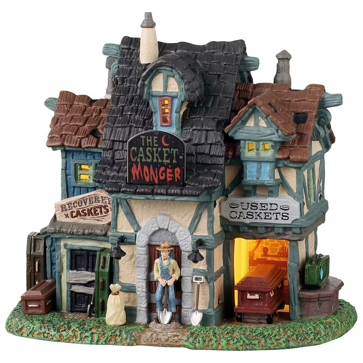 This is a Lemax Spooky Town building designed to look like an old-timey house turned into a casket store. There's a sign above the front door that reads "THE CASKET MONGER." There's a sign on the left side of the building that reads "RECOVERING CASKETS" and a sign on the right side that reads "USED CASKETS." There's a man shoveling near the front door and there are caskets everywhere.