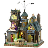 This is a Lemax Spooky Town building designed to look like a semi-rundown mansion. There's a sign that reads "SPELLS ABOUND." There are three bats flying above. There's a big wizard statue over one of the doors. You can see a wizard casting a spell on a cauldron, a person carrying books and a black cat on the sidewalk.