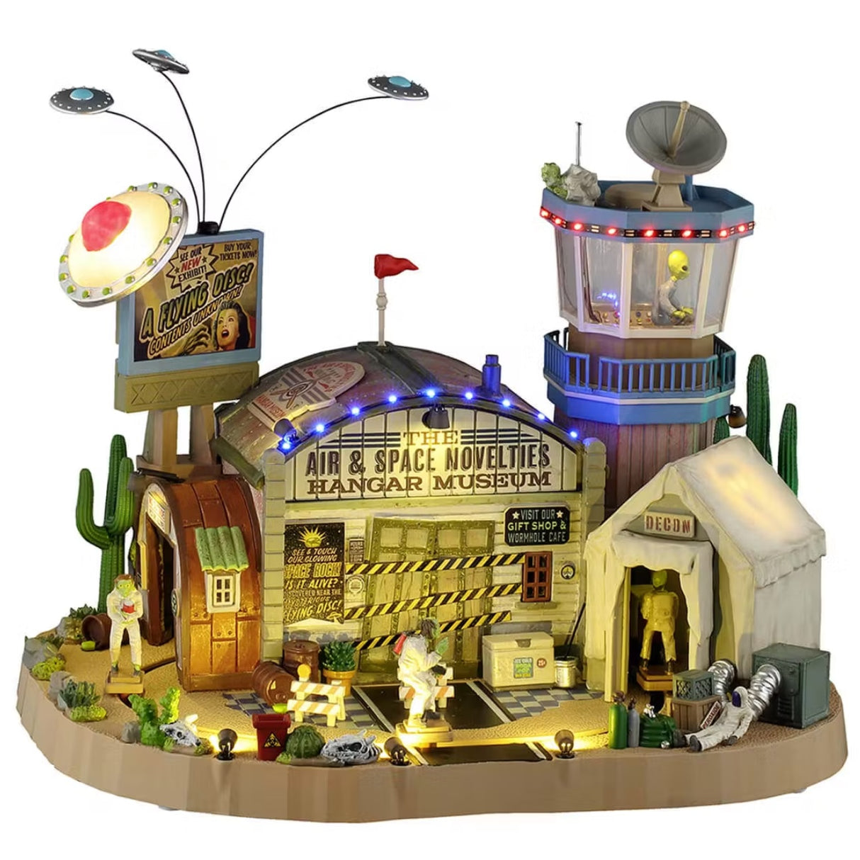 This is a Lemax Spooky Town building designed to look like a museum dedicated to aliens and UFOs. There are three circular UFOs flying above a sign that reads "See Our New Exhibit! A Flying Disc (Contents Unknown)." There are cactus plants, skulls, biohazard bins and barrels and boxes scattered everywhere outside. One of the signs on the building reads "See & Touch Our Glowing Space Rock. Is it alive?" The other sign on the building reads "Visit Our Gift Shop & Wormhole Cafe."