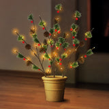 This is a lifestyle image showing the three branches staked into a flower pot on the floor inside a home. The warm white lights are on accenting the green pine needles, brown pine cones and red berries.