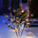 This is a lifestyle image showing the three branches staked into the snowy ground in front of a house. The warm white lights are on accenting the green pine needles, brown pine cones and red berries.